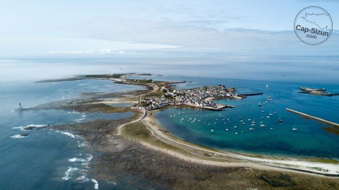Vue aérienne de l'île de Sein : le bourg serré autour de son port, et l'île basse qui s'étire à fleur d'eau vers le phare.